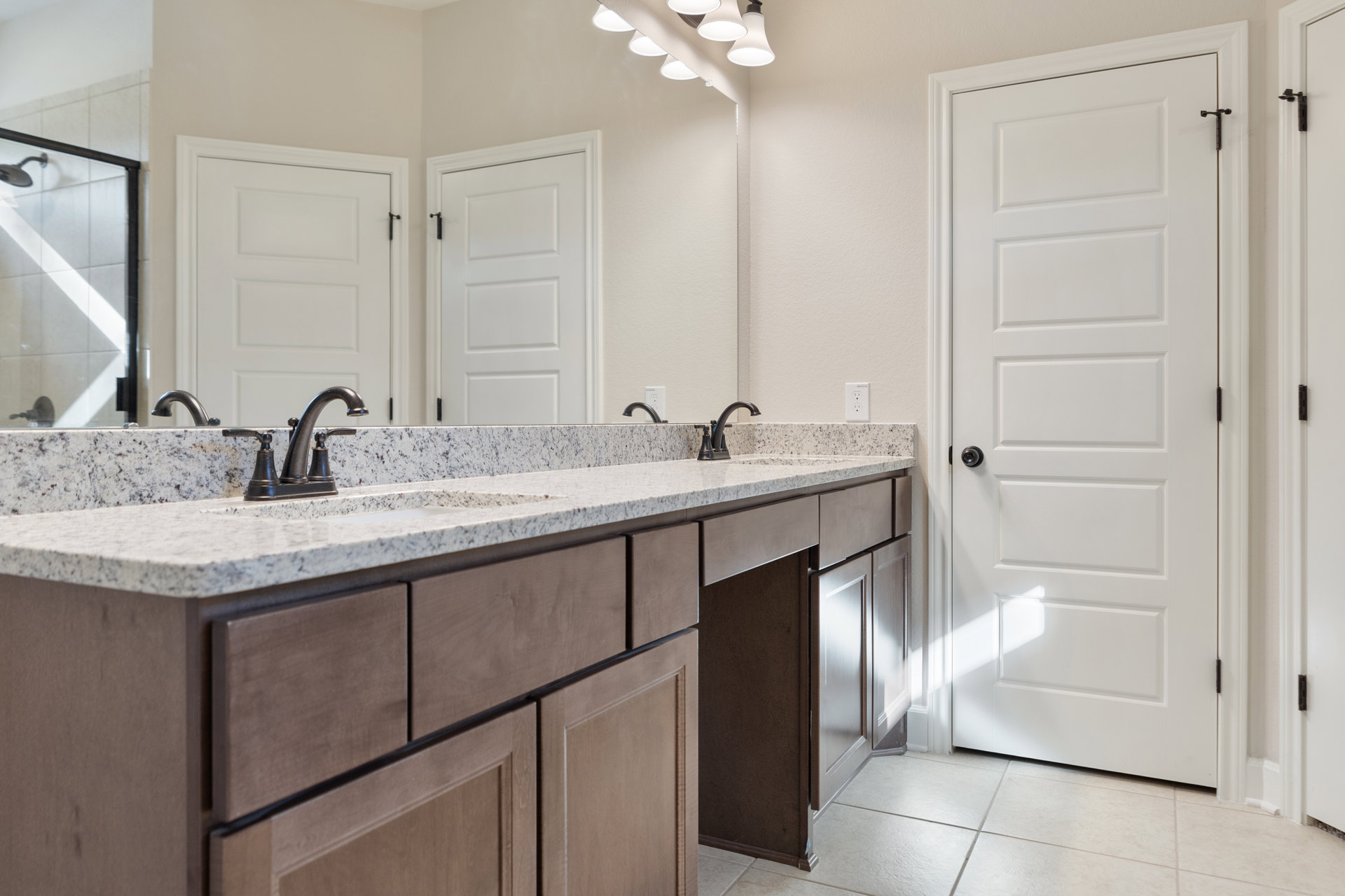 Double vanity bathroom with white cabinetry, black hardware, white tile wall, large mirror with lights, and multiple white doors