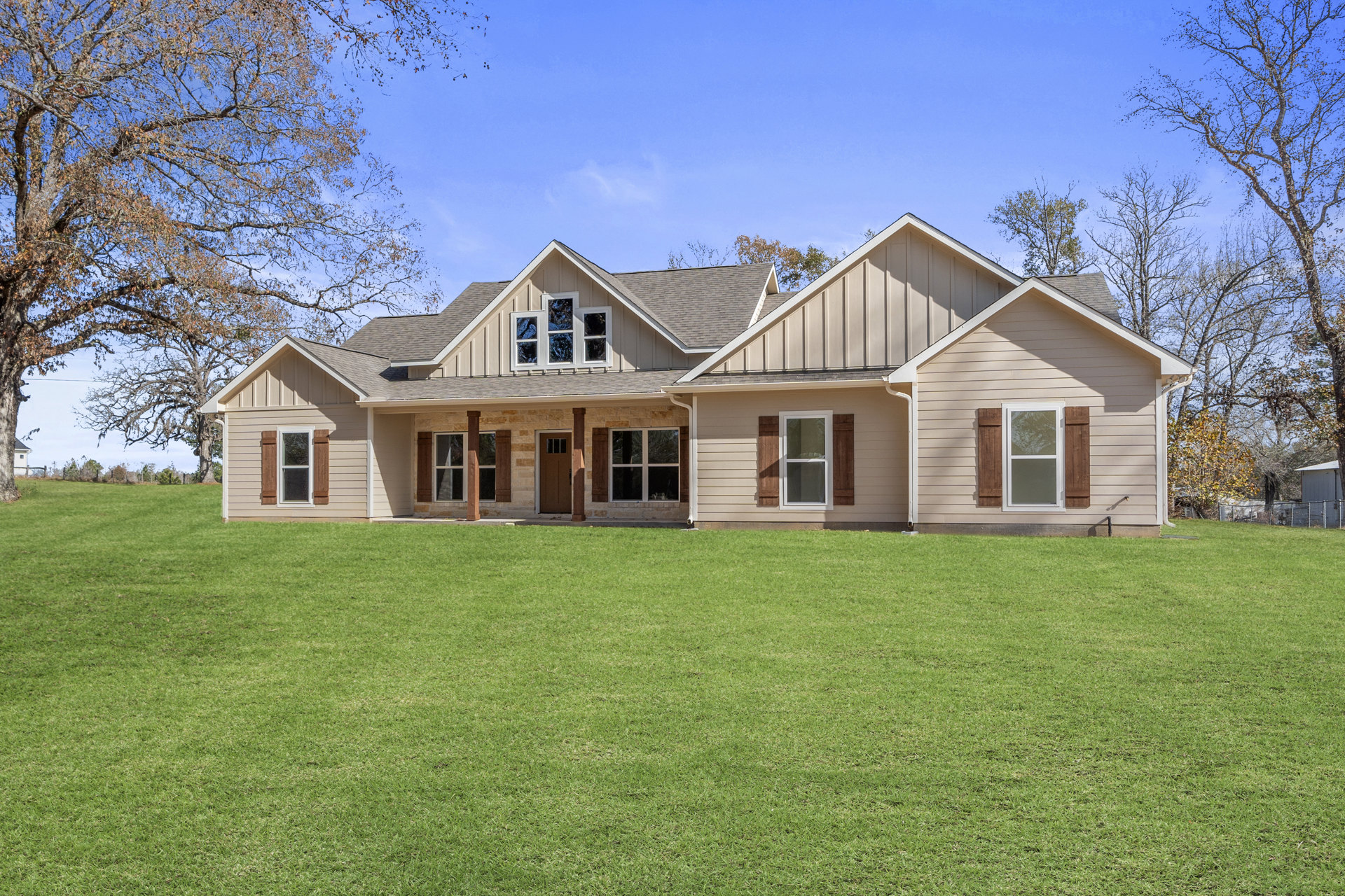 Two-story home with white-framed windows, gray shingle roof, covered porch, and green lawn bordered by trees; one window has broken glass.
