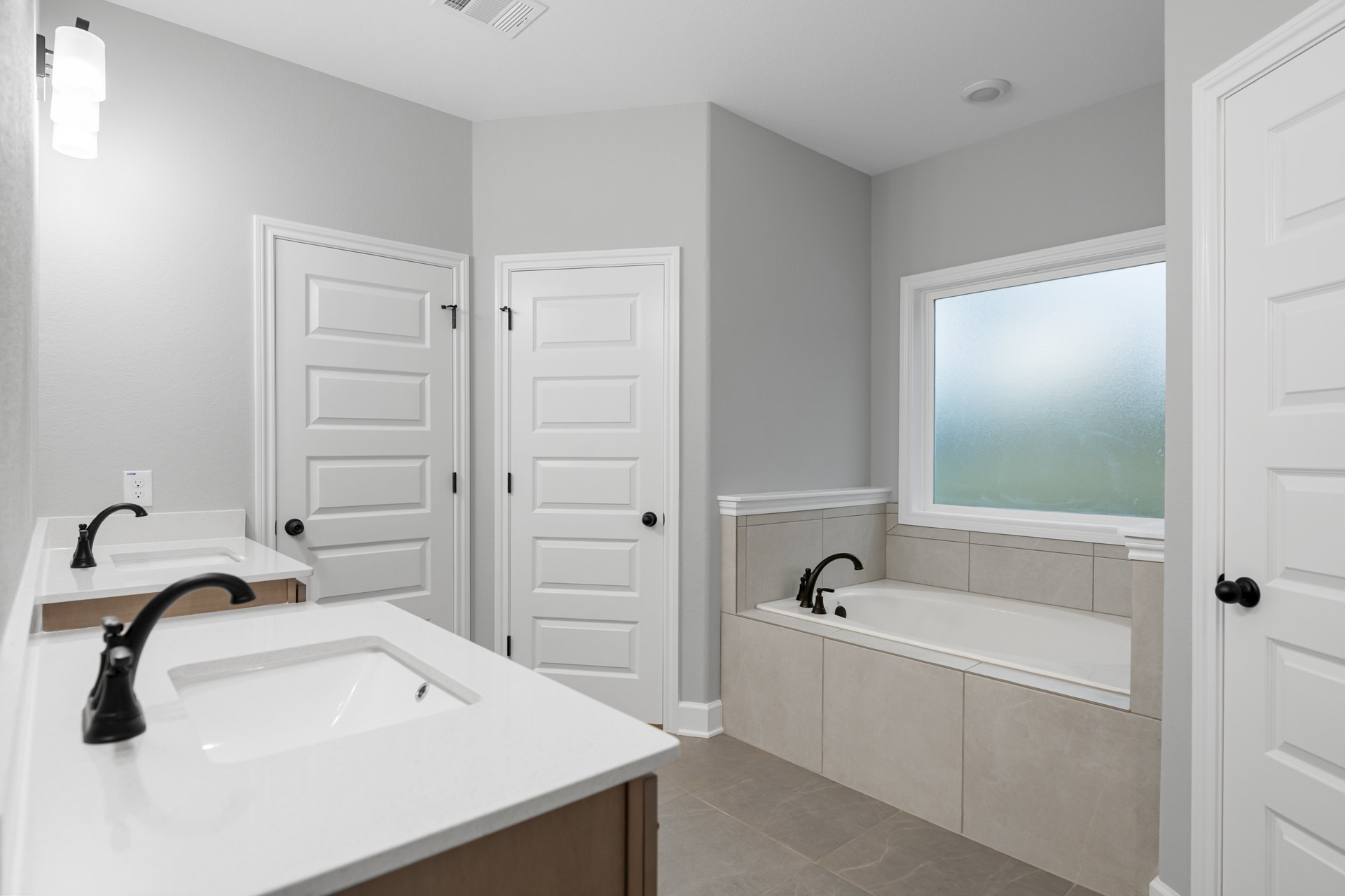 White freestanding tub beside a white vanity with black faucet, frosted glass window, white door with black handle, light tile flooring