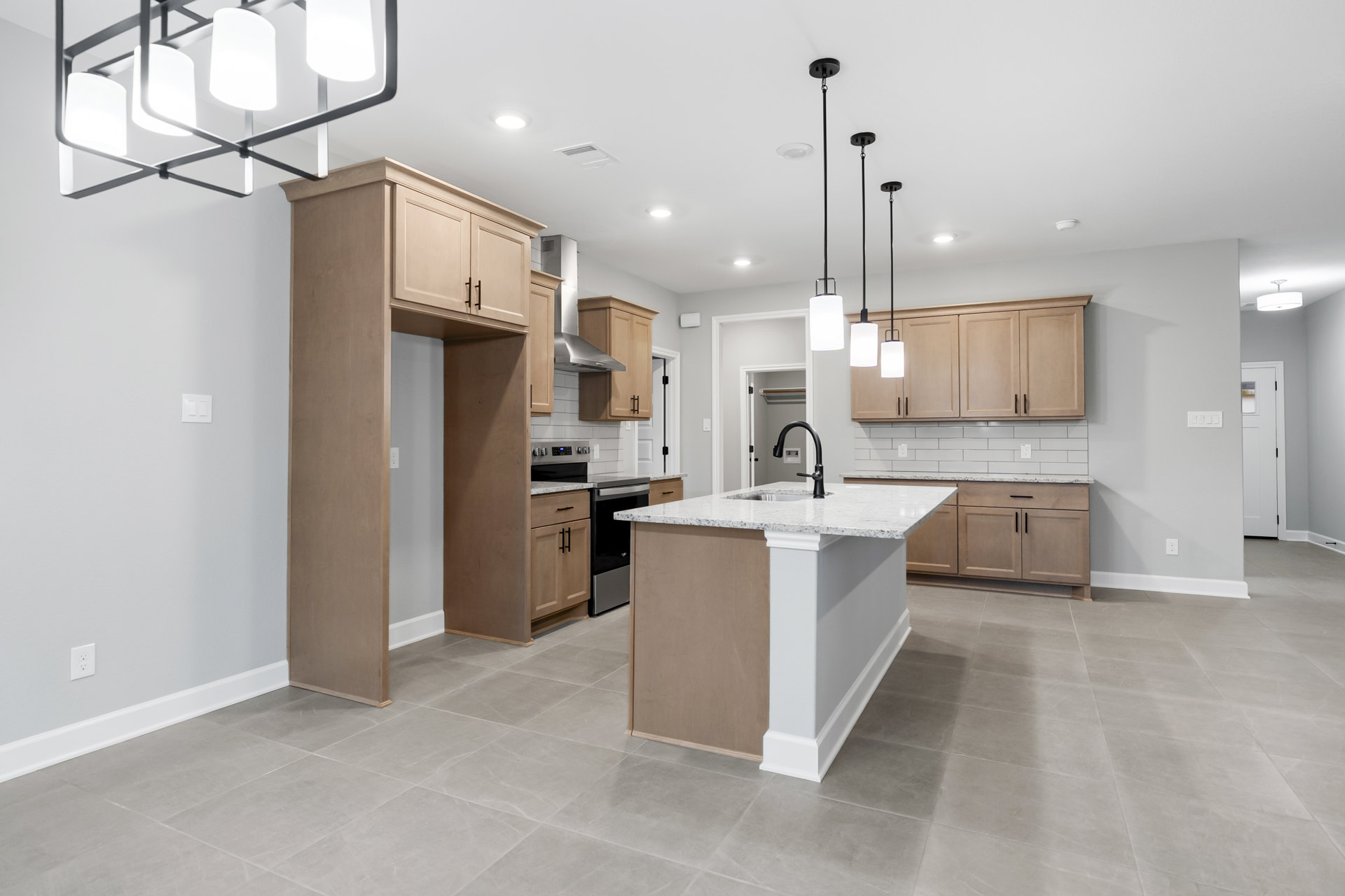 Open-concept kitchen featuring a large island with built-in sink, pendant light fixtures, white cabinetry, tiled backsplash, stainless steel stove, and white column accents.