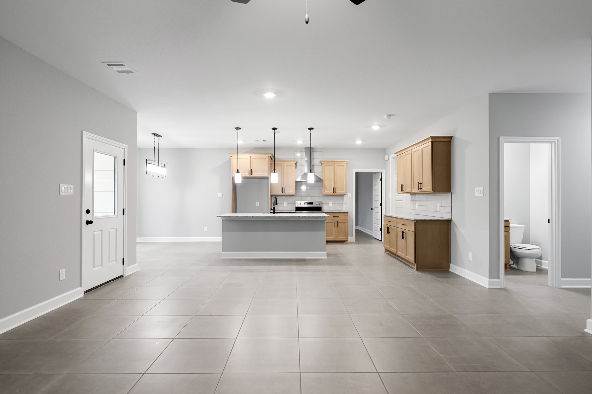 Open kitchen and dining area with tile flooring, wooden cabinets with black handles, ceiling fan, white door with window, kitchen counter with sink, and adjacent bathroom with