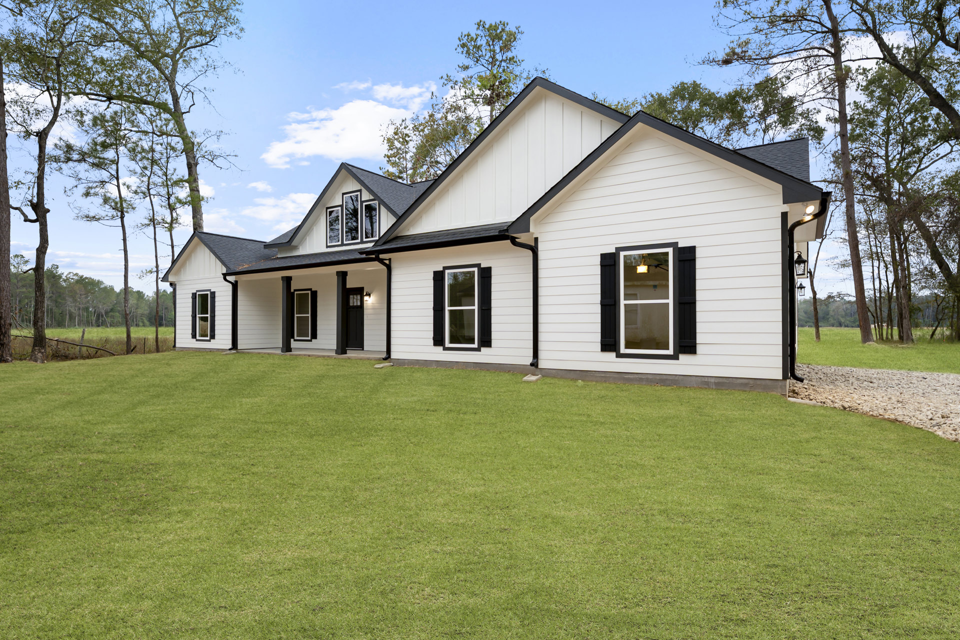 White siding house with black trim and shutters, illuminated window, green lawn, mature tree in background, wooded surroundings.