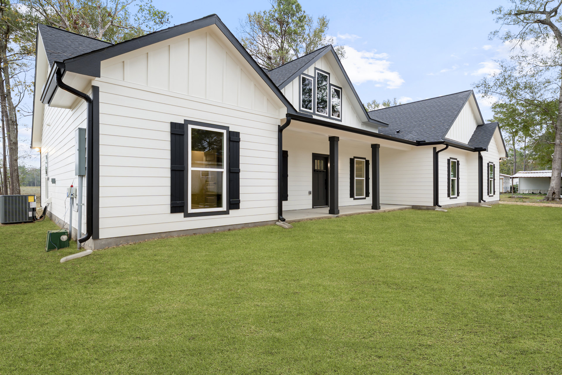 White siding house with black trim, white-framed windows reflecting trees, manicured lawn, green utility box with round light, large gray HVAC unit with black hose on side