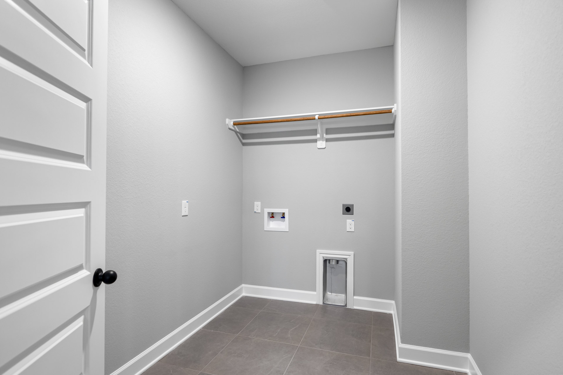 White-walled room with a small closet, black door, grey tile flooring, and visible plumbing valves on a white box