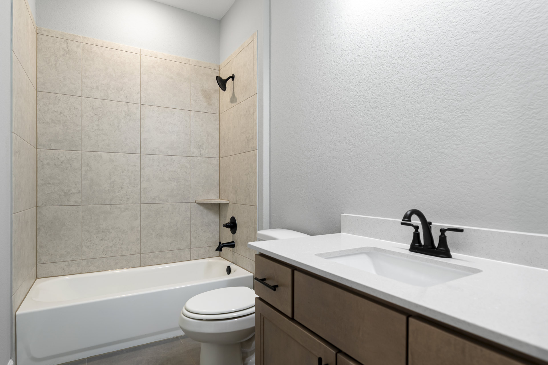 Modern bathroom featuring a white bathtub beside a toilet with lid up, wall-mounted shower head on light tile, and a sink with chrome faucet atop a wood cabinet.