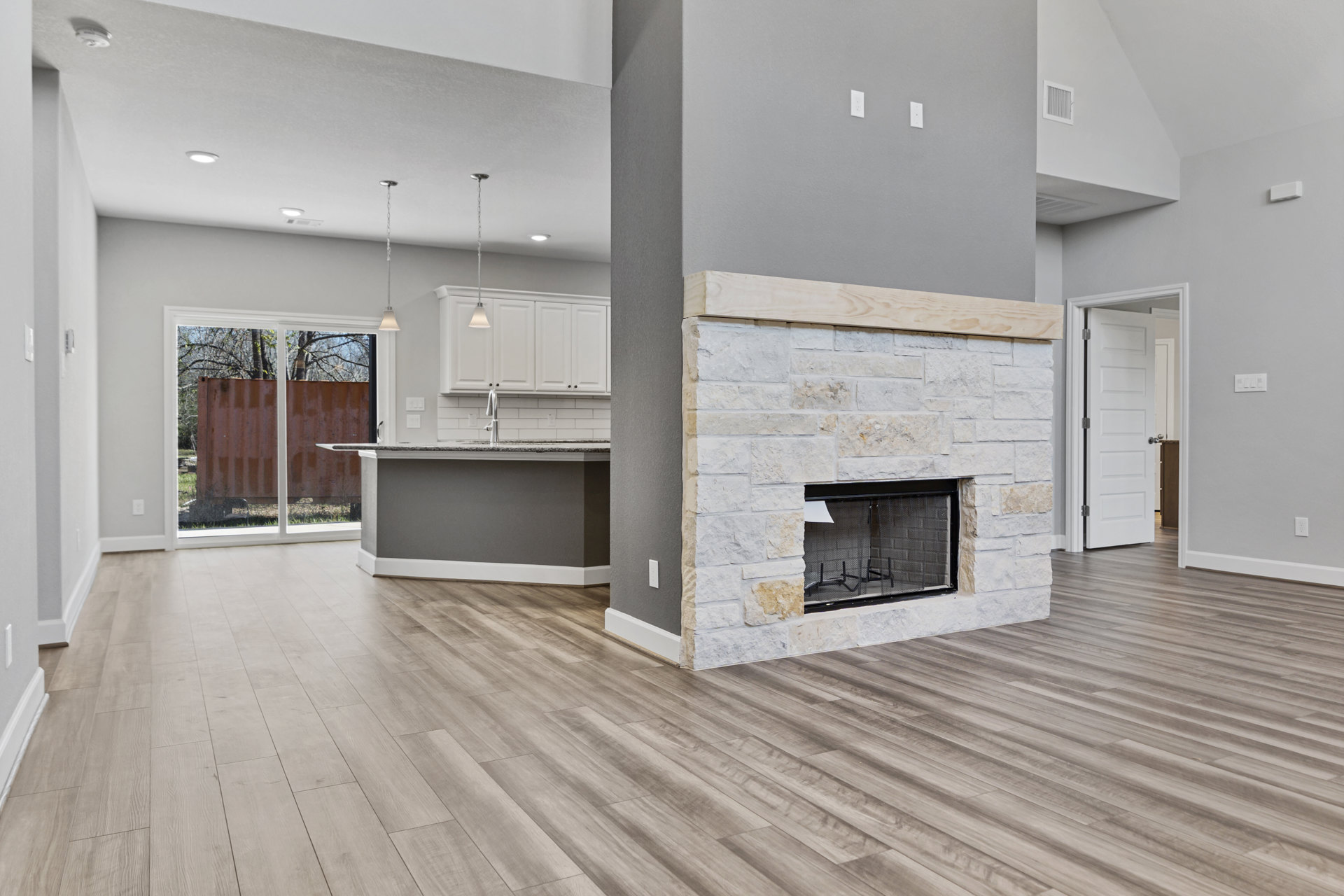 Living room with wood flooring, central fireplace framed in wood, fireplace screen with paper, open kitchen area, white door with silver knobs, large window and container in
