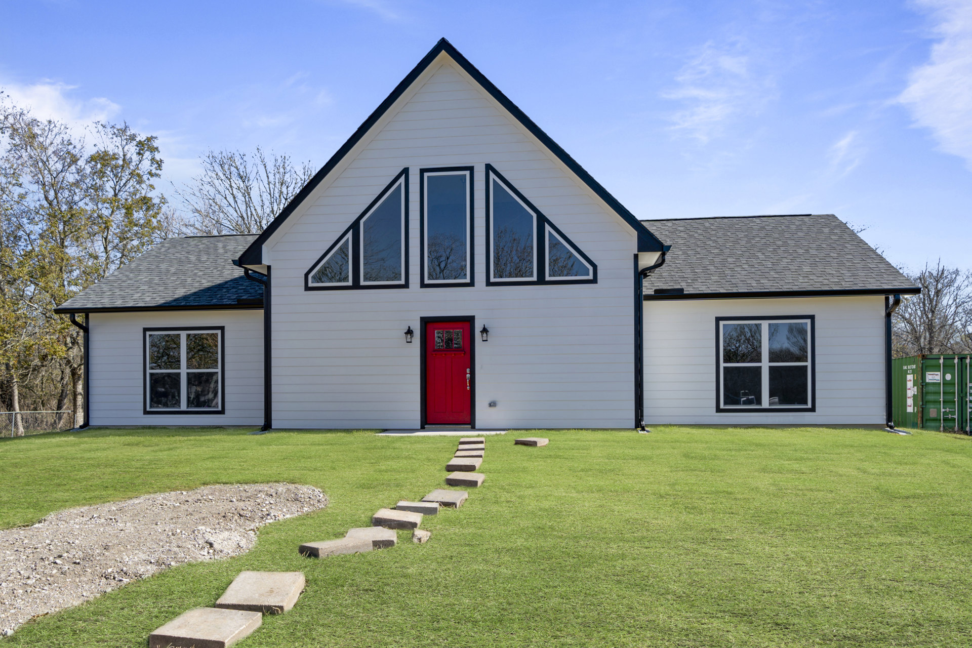 White cottage-style house with red front door, white-framed windows reflecting trees, green grass lawn, and cloudy sky