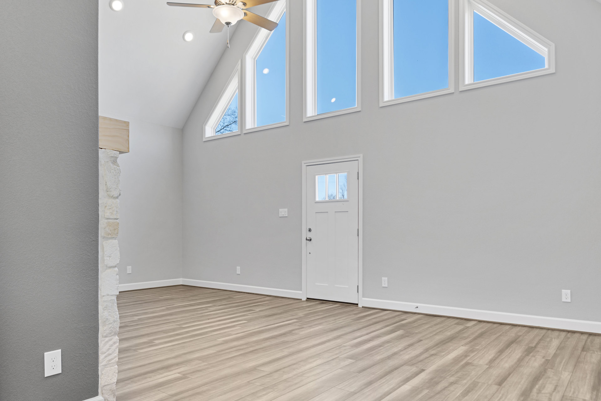 White paneled door with glass window, wood laminate flooring, white plaster walls, ceiling fan with light fixture, rectangular window showing blue sky.