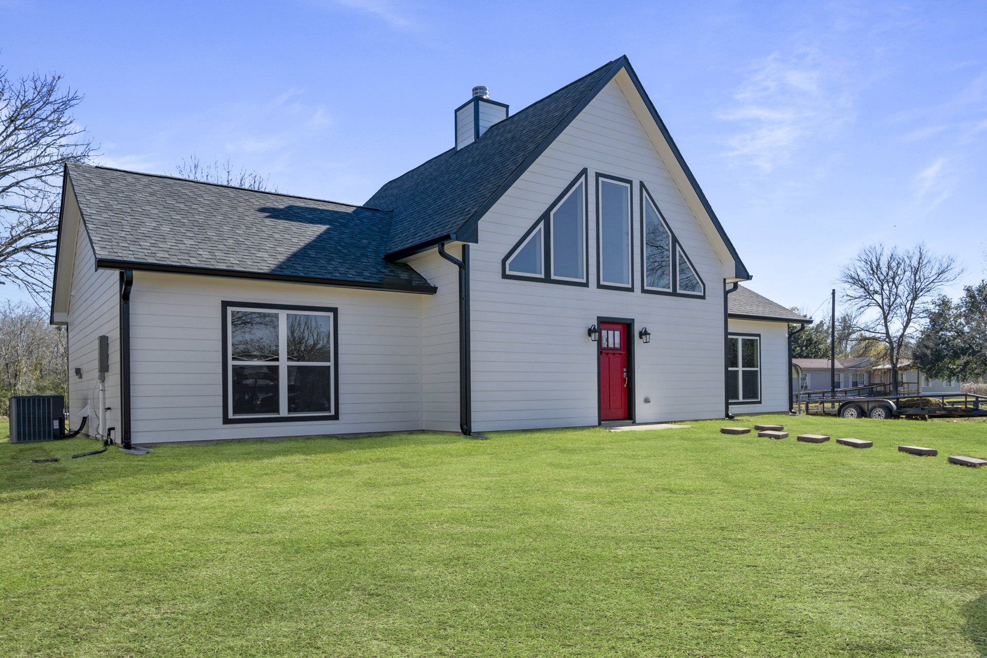 White farmhouse with black roof, red front door with glass window, large windows reflecting trees, green lawn, cloudy sky, and mature trees in background
