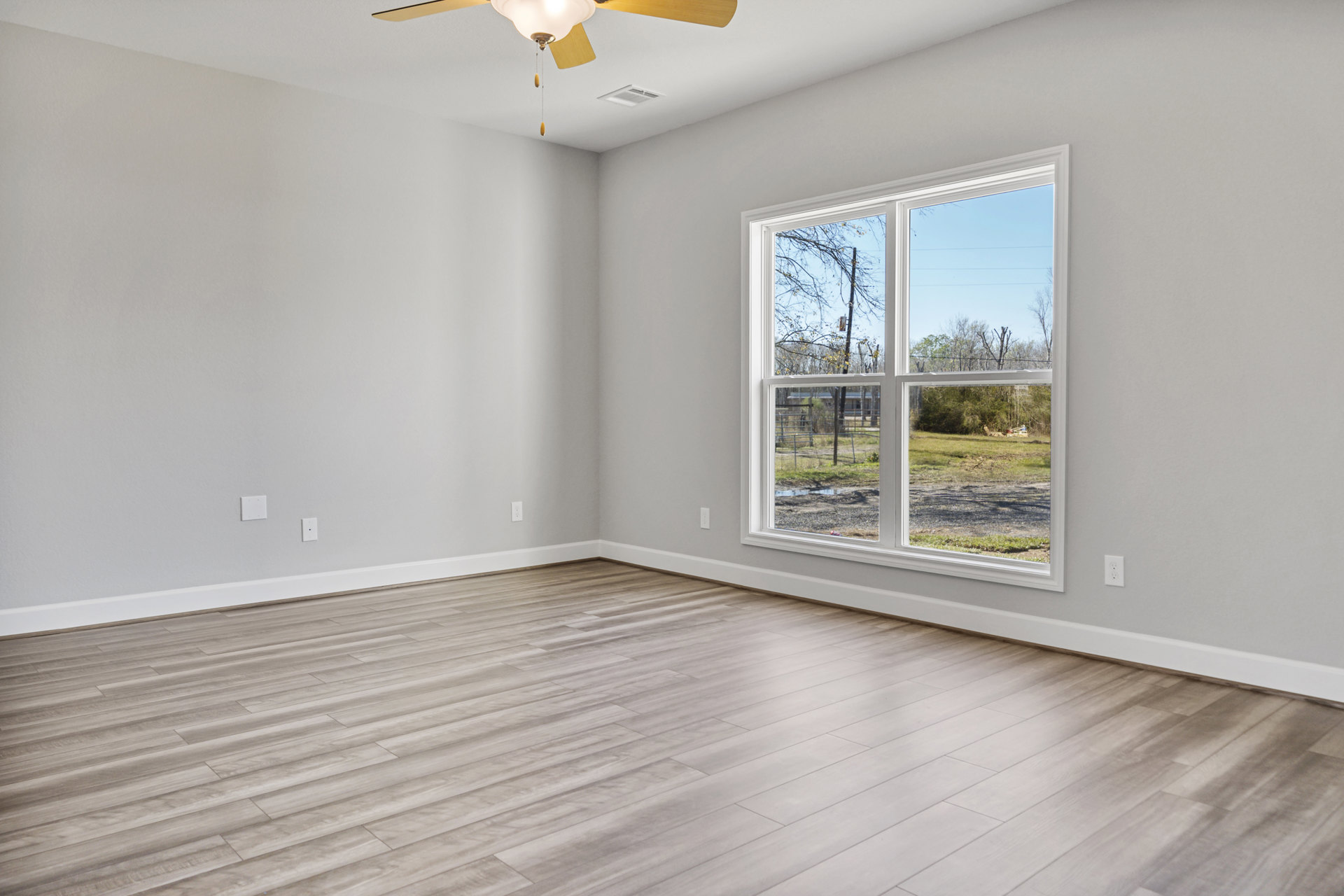 Wood flooring room with white walls, ceiling fan with light fixture, large window overlooking green yard and trees, ceiling vent visible