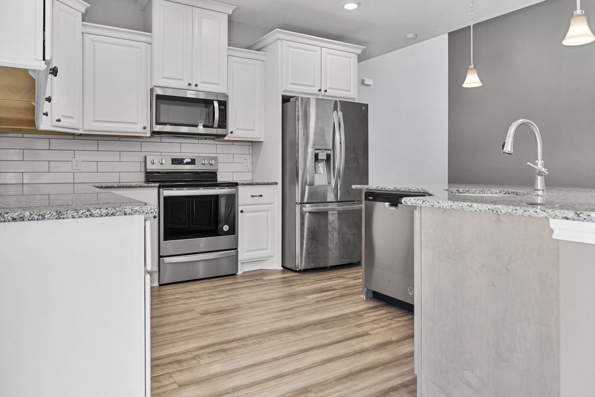 Kitchen with stainless steel refrigerator, oven, and microwave, white cabinetry, silver faucet with black handle, wood flooring, and light countertops.