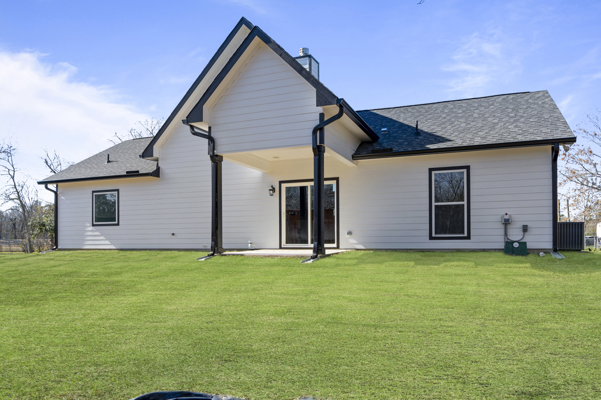 White house with black gutters, large windows, glass door with black trim, green lawn featuring a shovel, tree visible outside window, partly cloudy sky overhead