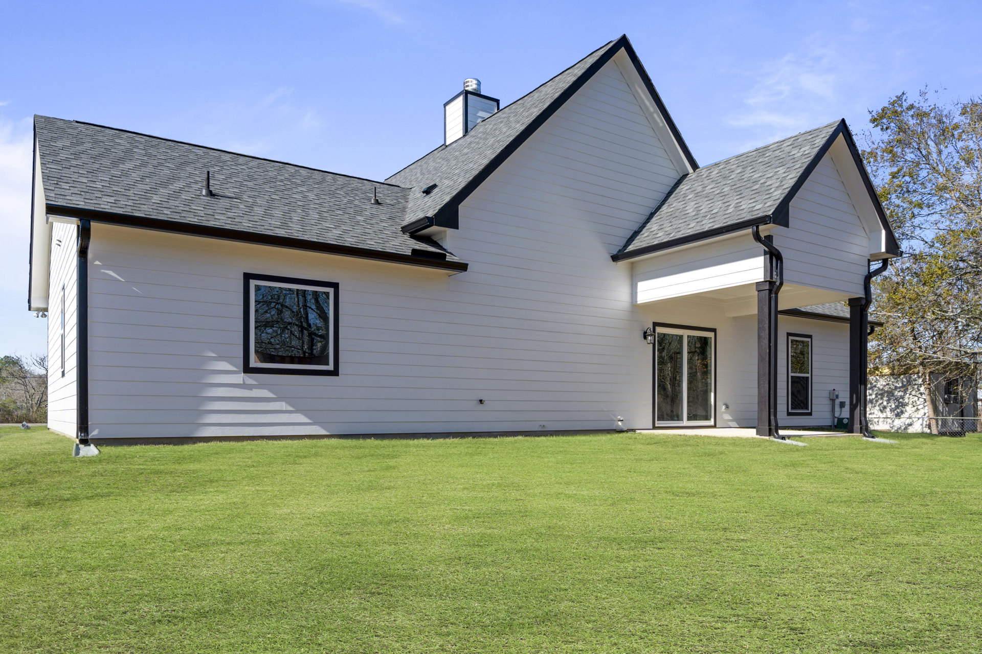 White siding house with black roof, illuminated window, green lawn in front, mature trees in background, clear sky overhead