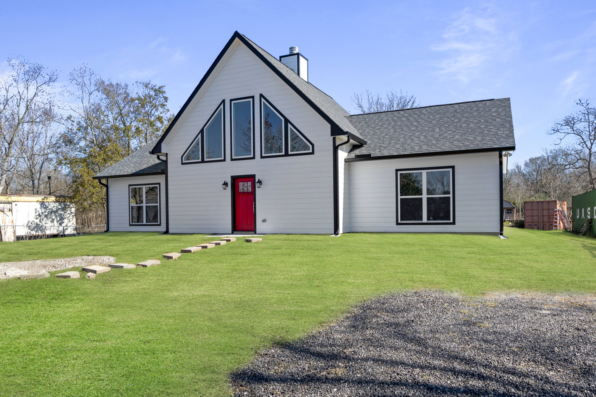 White house exterior with red door featuring glass panes, white-framed window, green grass lawn, trees in background, partly cloudy sky