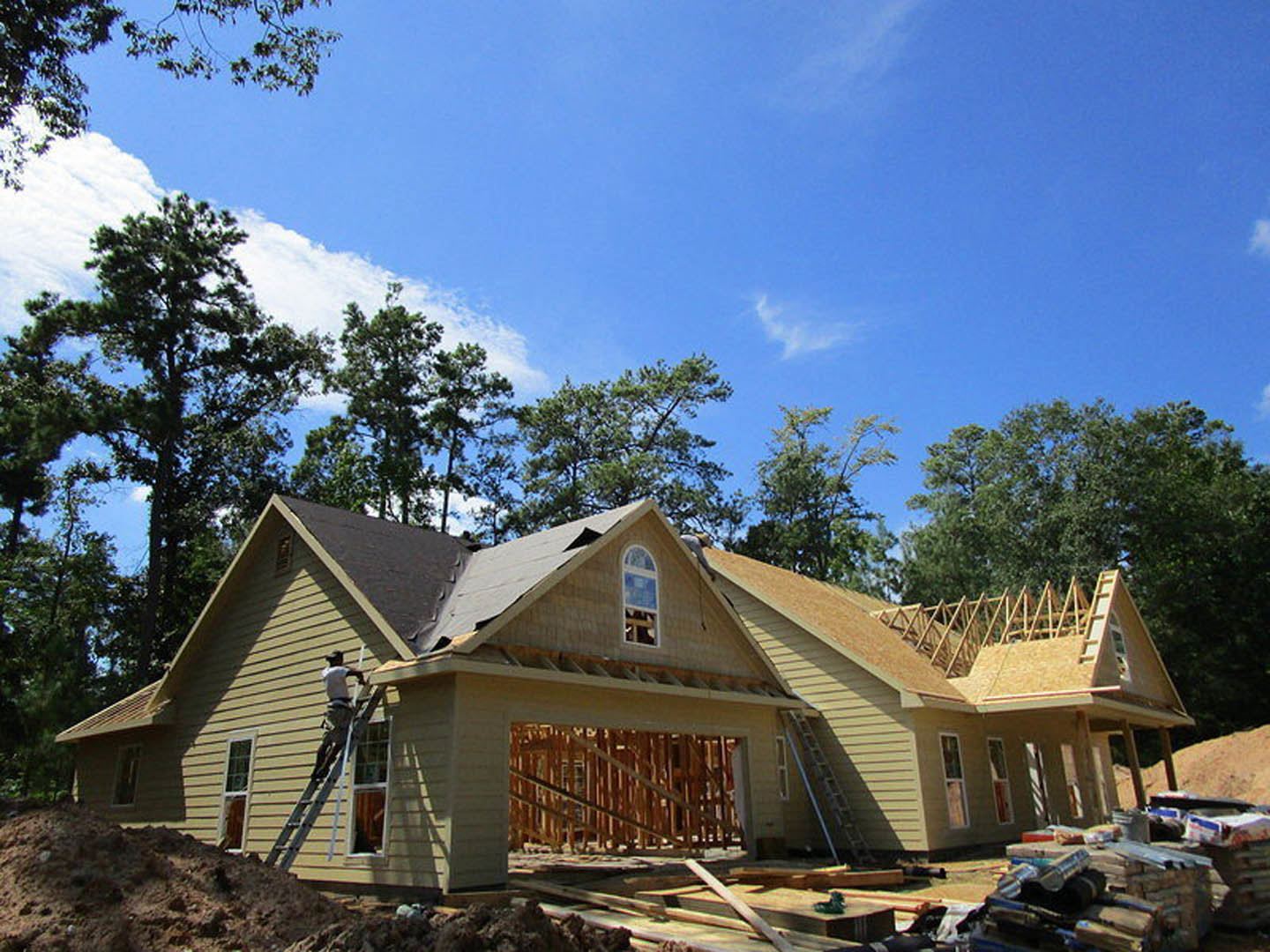 Wood-framed house under construction with white window frames, ladder leaning against exterior, surrounded by trees and cloudy sky