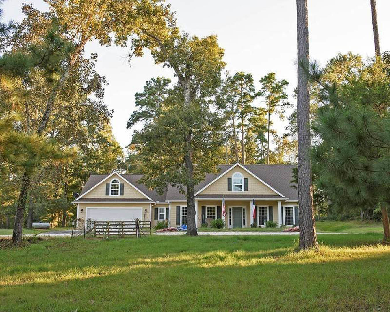 White farmhouse with covered front porch, large windows, and a fenced yard featuring mature trees and manicured grass