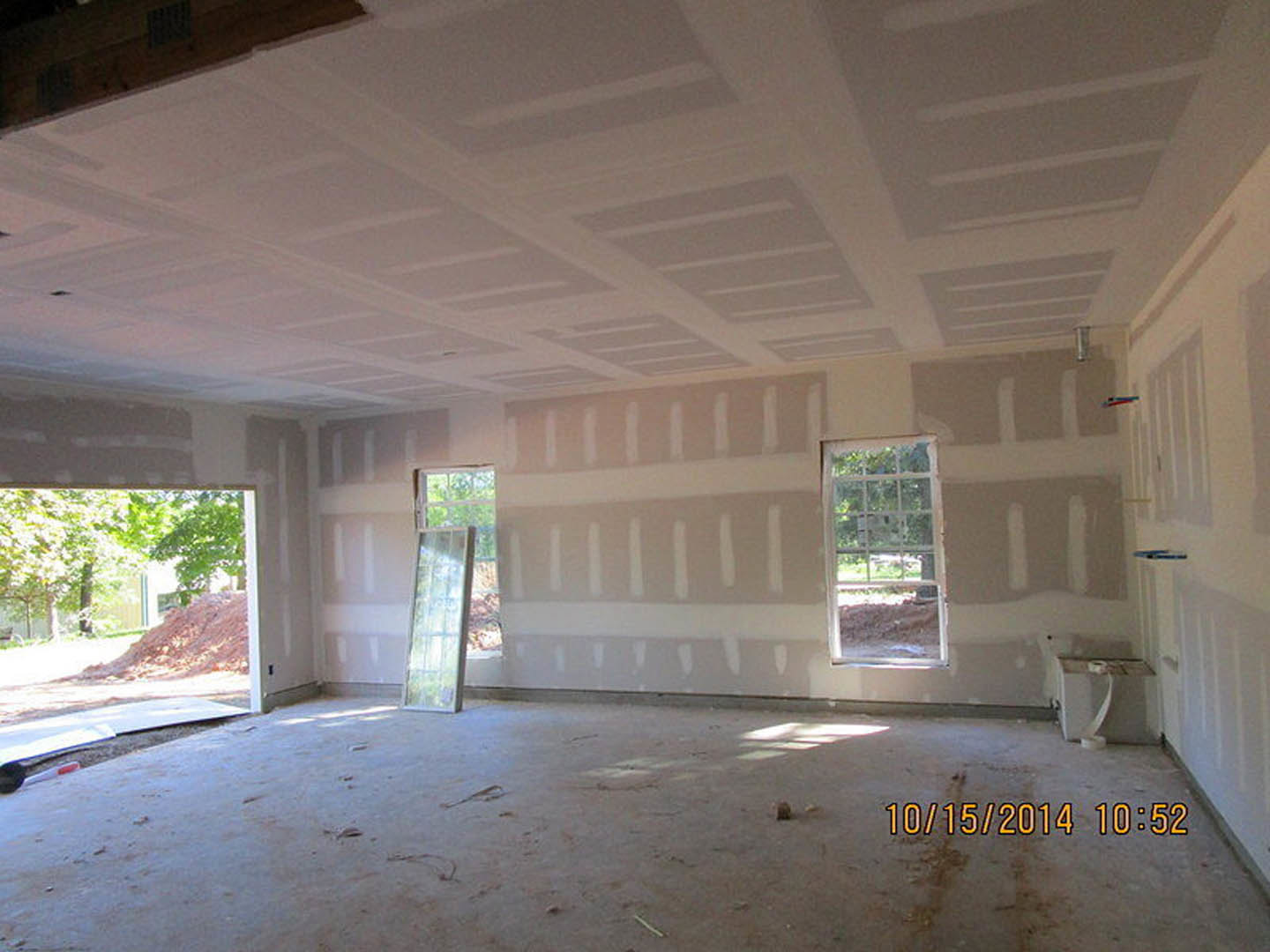 Bright room with white plaster walls, large windows letting in daylight, white ceiling with subtle grey lines, and a reflective mirror; floor shows visible dirt.