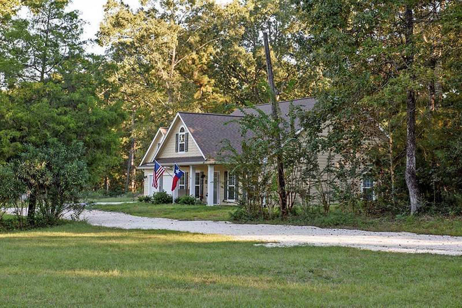 White farmhouse with gabled roof, American flag on front porch, mature trees in background, manicured lawn, gravel driveway
