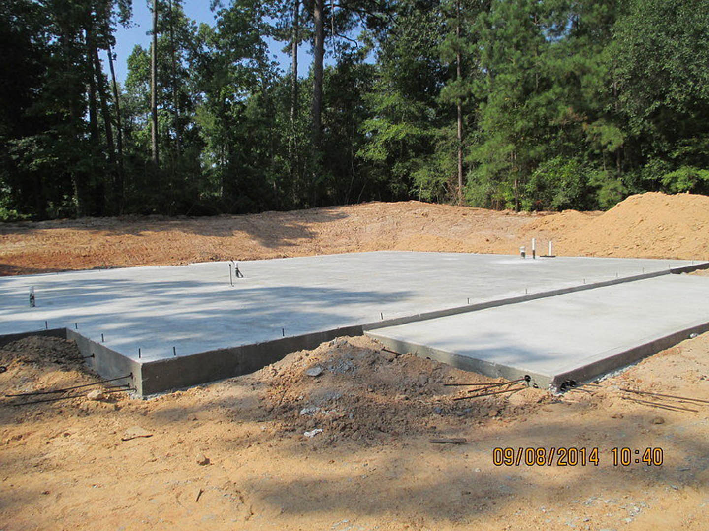 Freshly poured concrete slab surrounded by piles of dirt, bordered by a dirt road and trees in the background, with construction materials visible on site