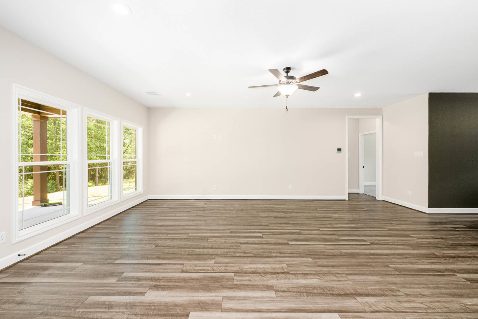 Open living space with wood flooring, white plaster walls, ceiling fan with light fixture, row of windows framed by wooden posts, and a black accent wall.