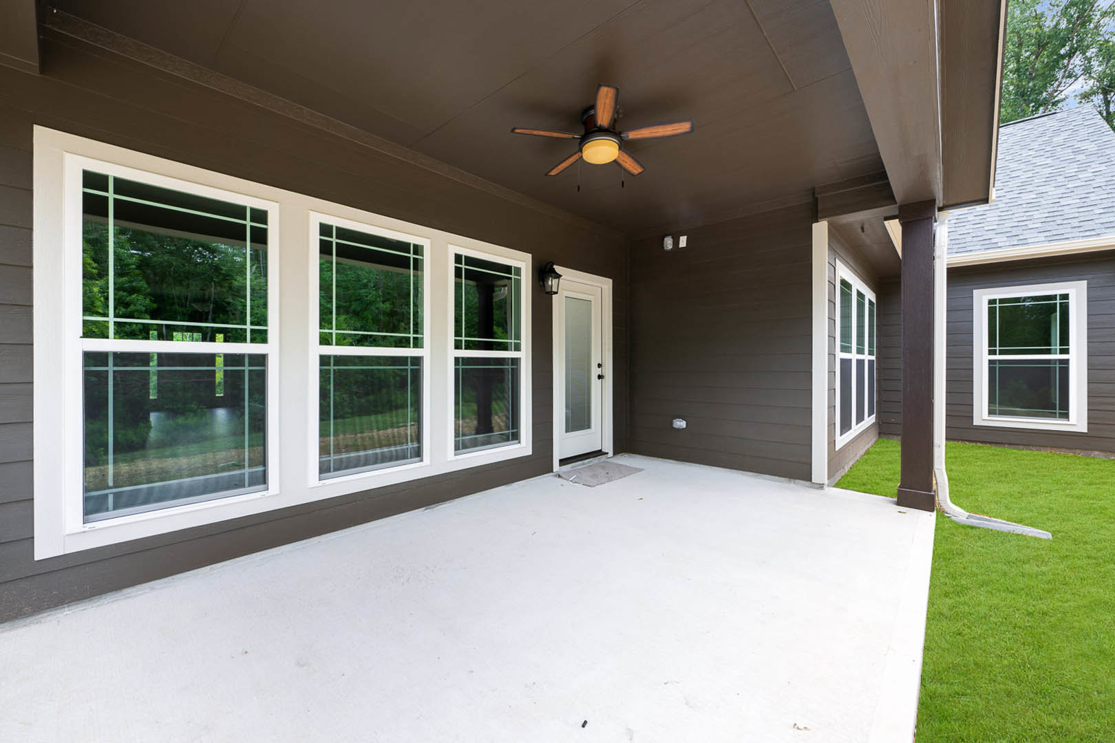 Covered porch with white floor, ceiling fan with light, row of windows and door, green lawn bordered by trees visible through windows