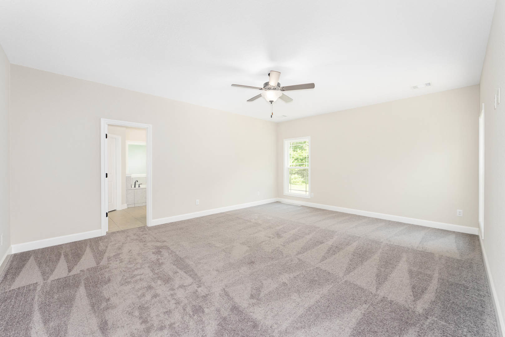 Carpeted bedroom with white walls, ceiling fan with light fixture, large window with white trim, partial view of bathroom featuring sink and faucet