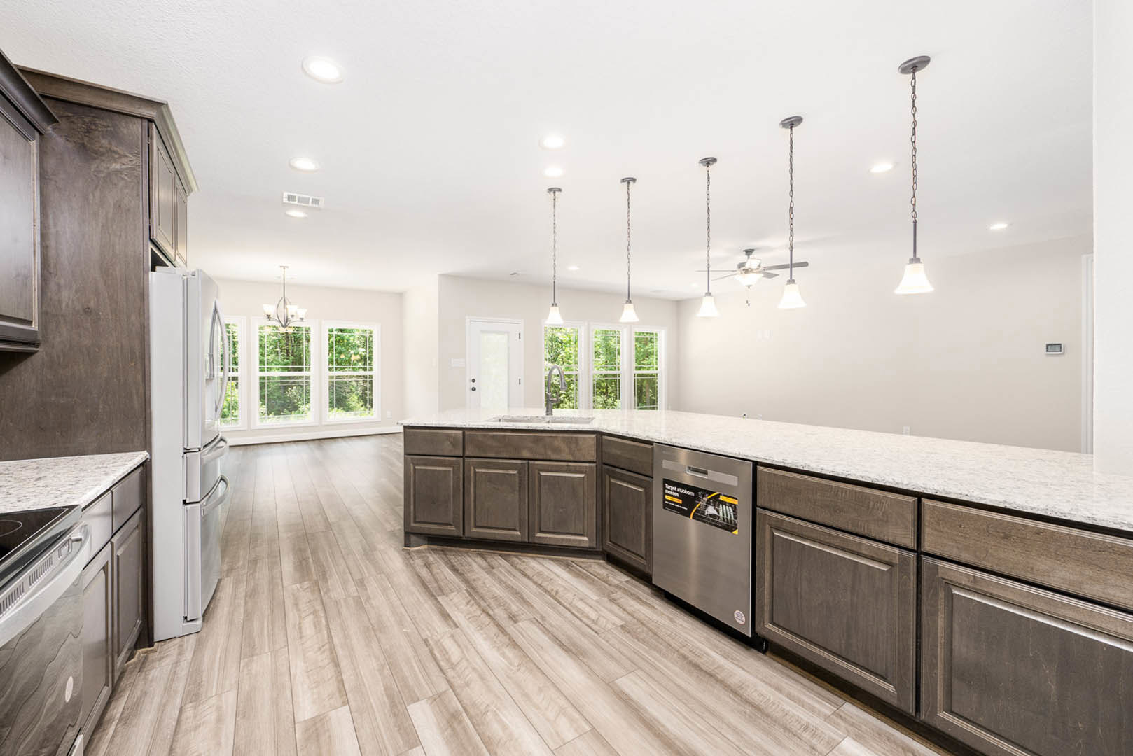 Spacious kitchen featuring a large central island with stone countertop, stainless steel refrigerator, white cabinetry, light wood flooring, and a window overlooking green trees.