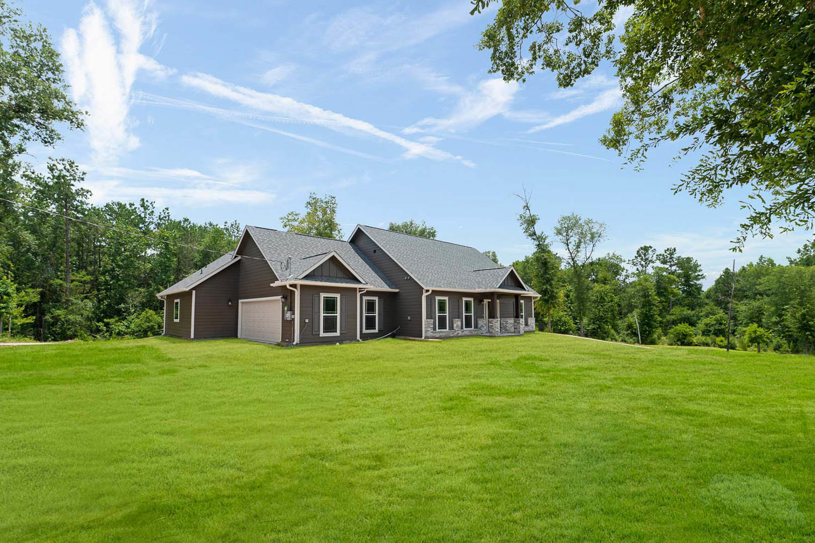 Brown siding house with white-framed windows and white garage door, expansive green lawn in foreground, blue sky with scattered clouds above