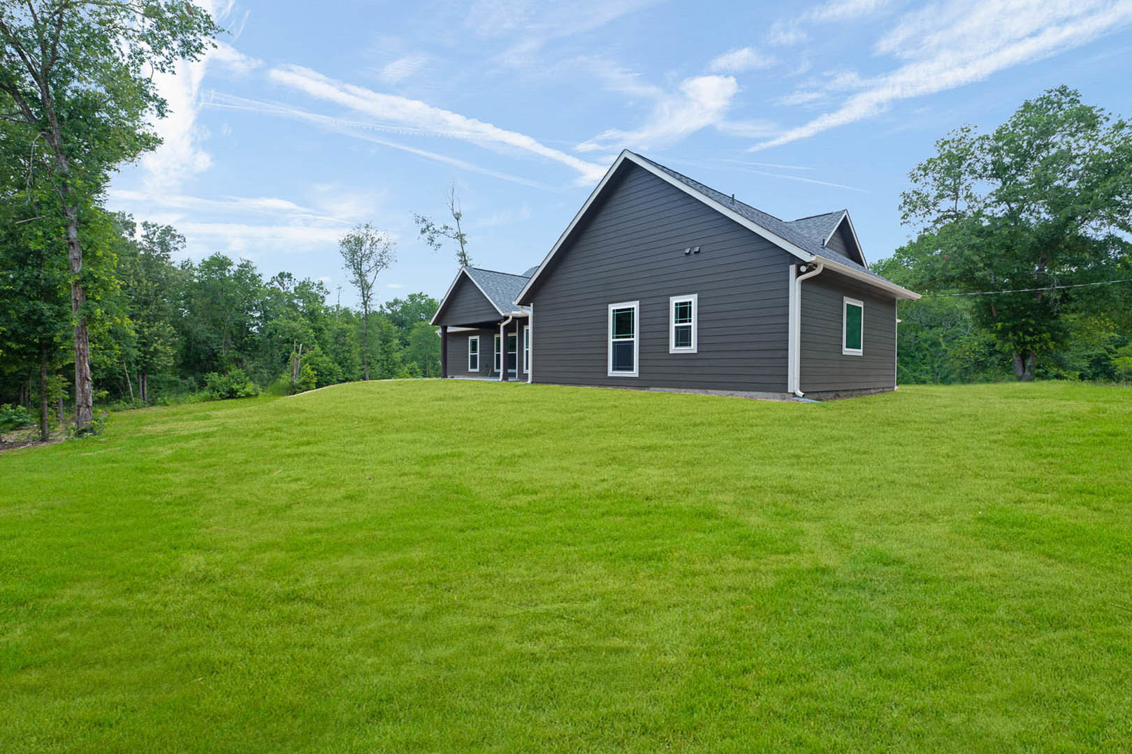 White farmhouse with large windows and a covered porch, set atop a green grassy hill with mature trees and blue sky in the background