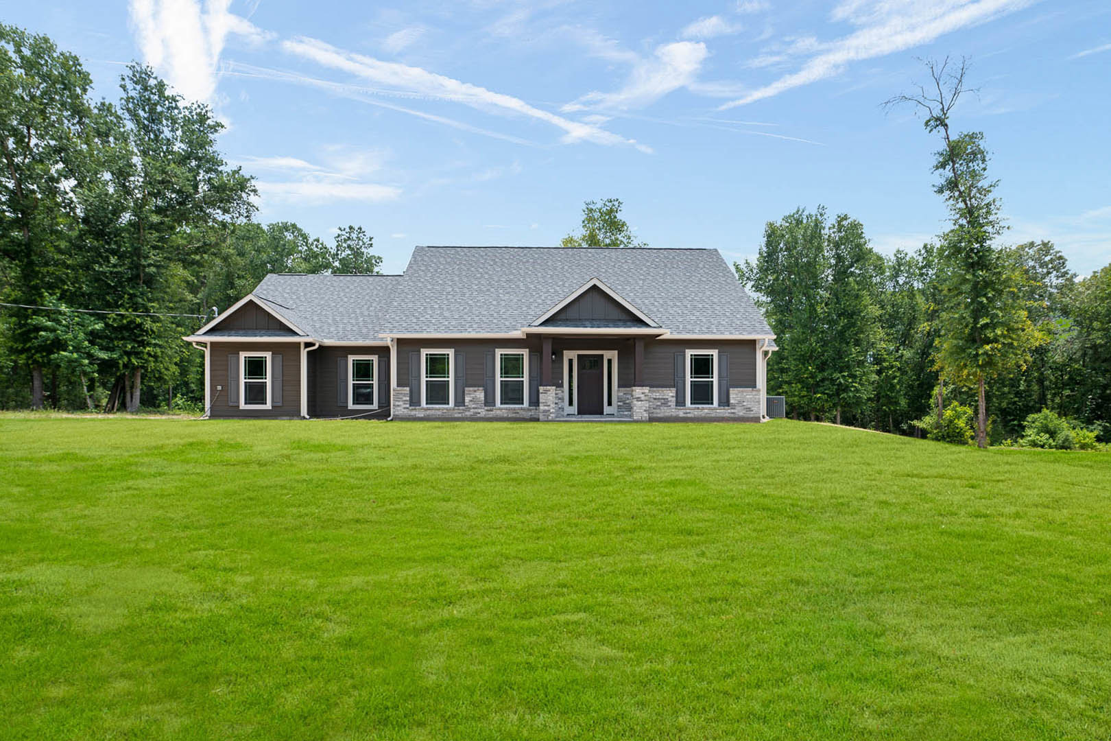 Brick farmhouse with grey roof, expansive green lawn, white-framed windows, black front door with white trim, mature trees, and partly cloudy sky