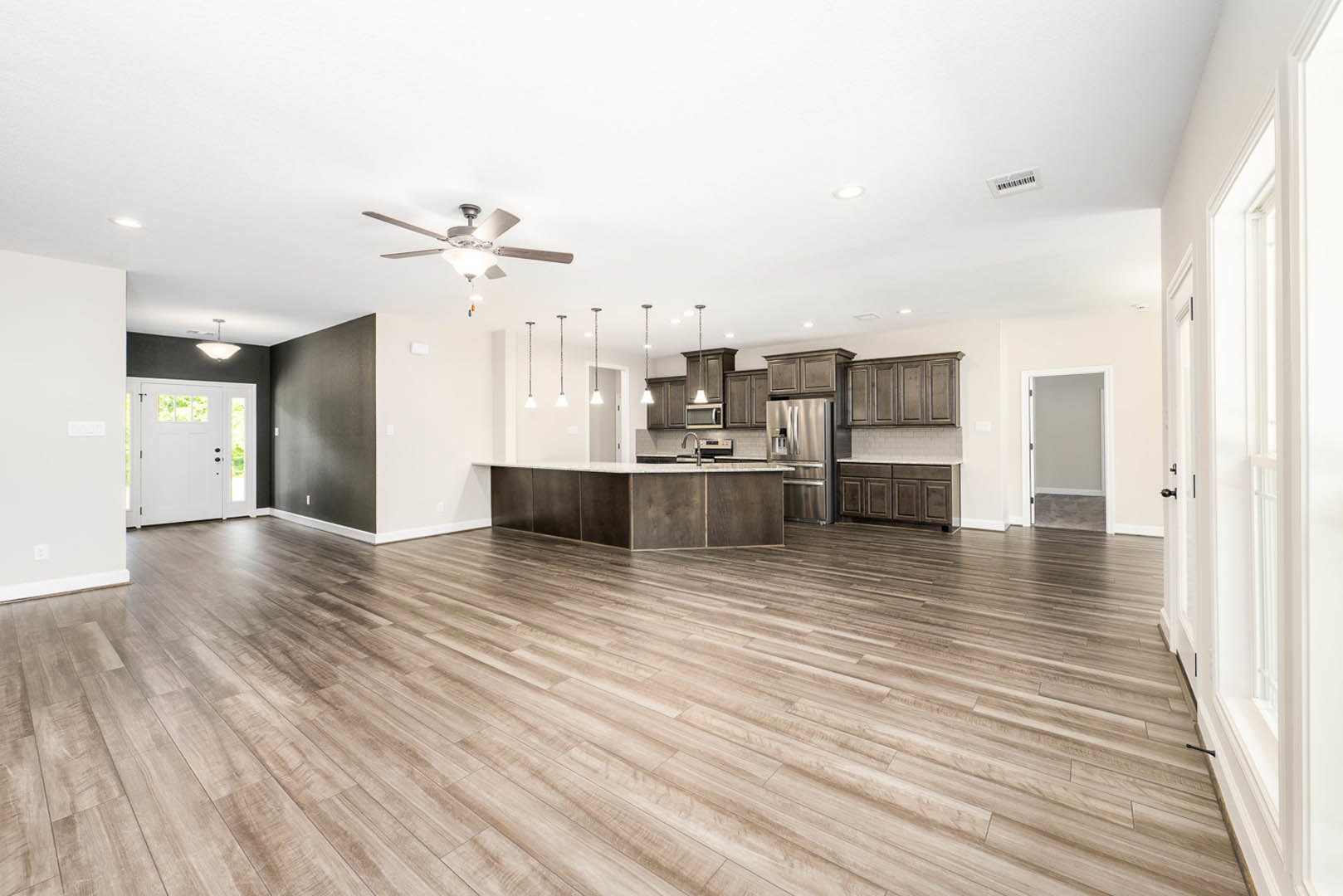 Spacious open floor plan featuring hardwood flooring, modern kitchen with light countertops, ceiling fan with light fixture, white framed door with glass panels, and partial view