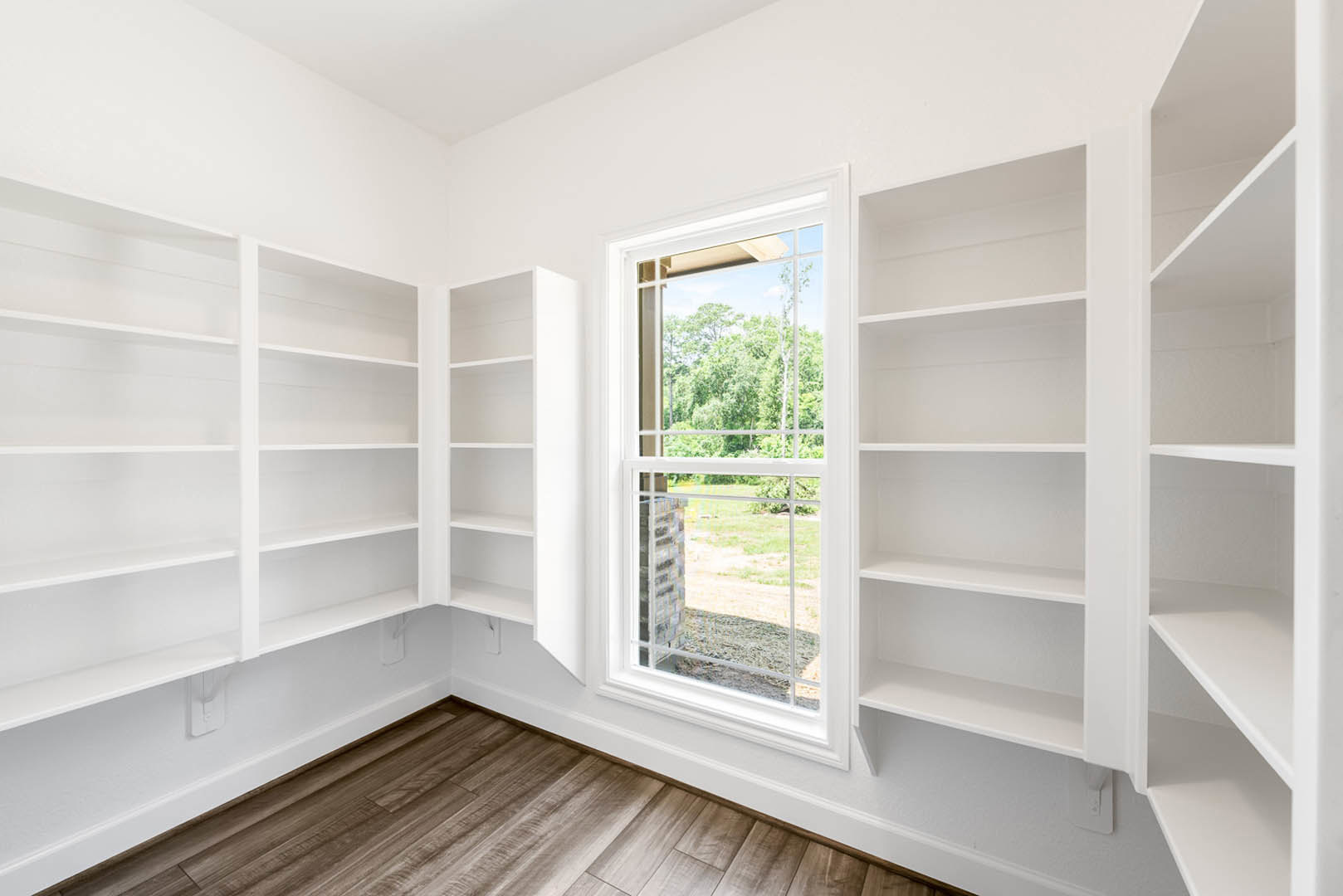 White built-in shelves along a plaster wall, wide window overlooking grassy field and trees, hardwood floor, minimalist decor