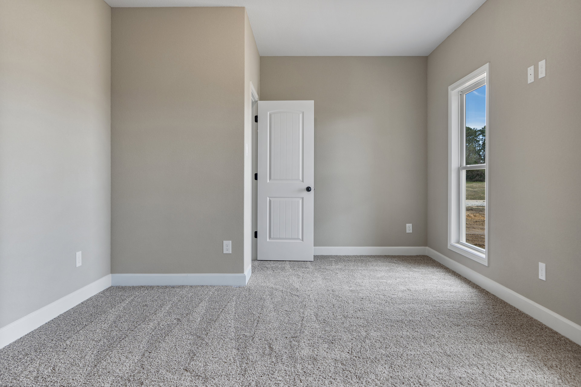 Carpeted room with white walls, white door featuring black knob, window showing trees outside, white wall with black fixture, white-framed window