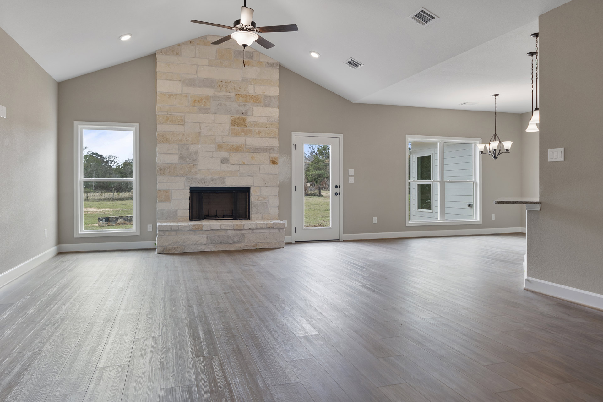 Spacious living room featuring wood flooring, brick fireplace with stone surround, ceiling fan with light fixture, large window overlooking trees, and white door opening to yard
