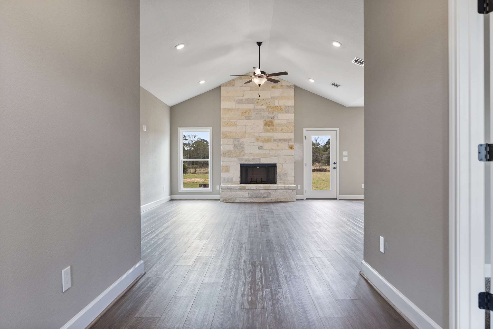 Living room with brick fireplace, ceiling fan with light fixture, large window and door offering views of trees and field, neutral plaster walls, wood flooring, black curtain