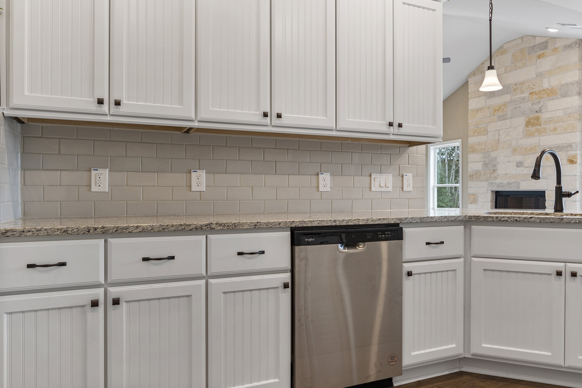 White shaker cabinets, stainless steel dishwasher with handle, quartz countertop, white subway tile backsplash, window overlooking trees, close-up of electrical outlet on wall