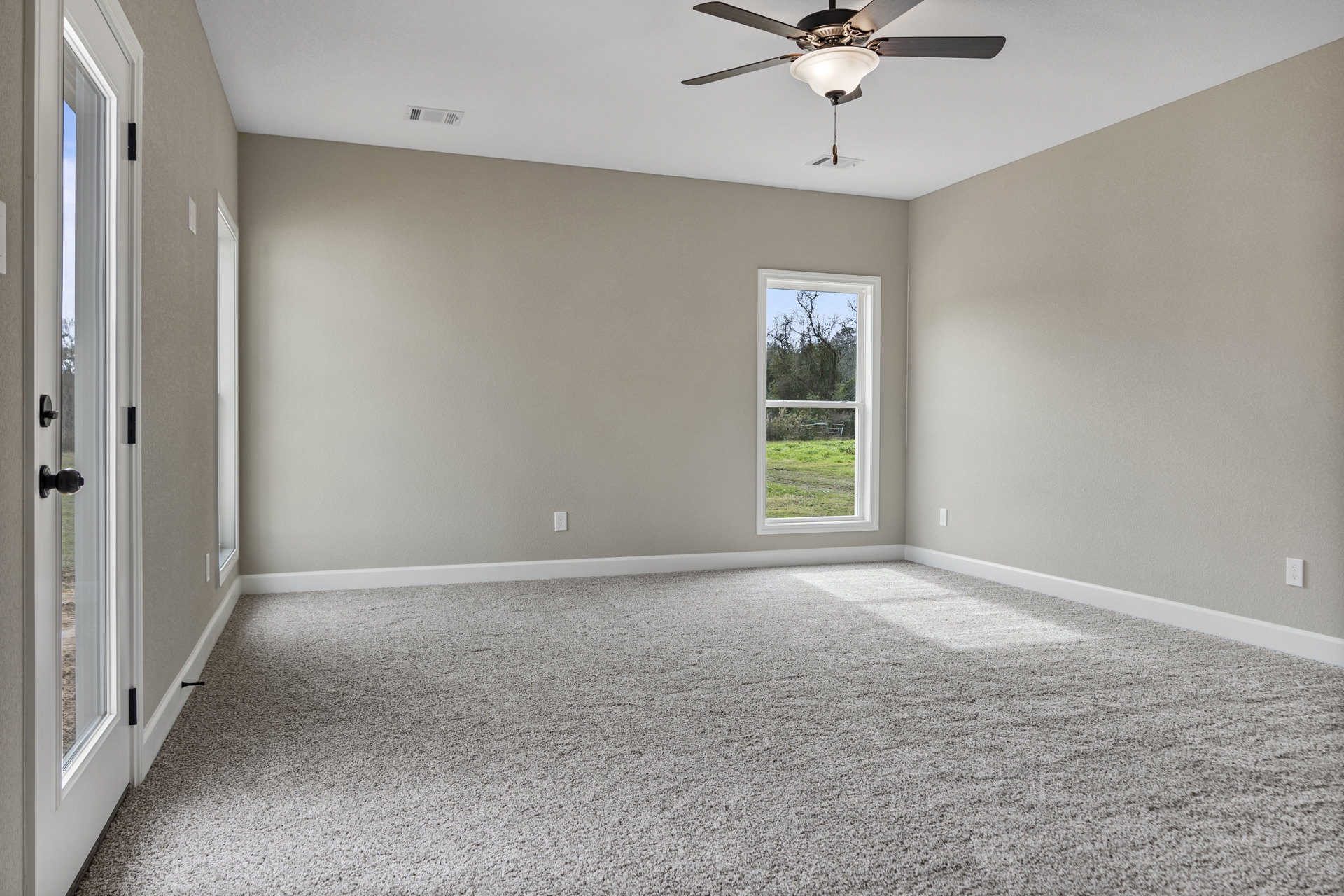 Carpeted bedroom with white walls, ceiling fan with light fixture, large window overlooking grassy field and trees, and paneled door with metal doorknob