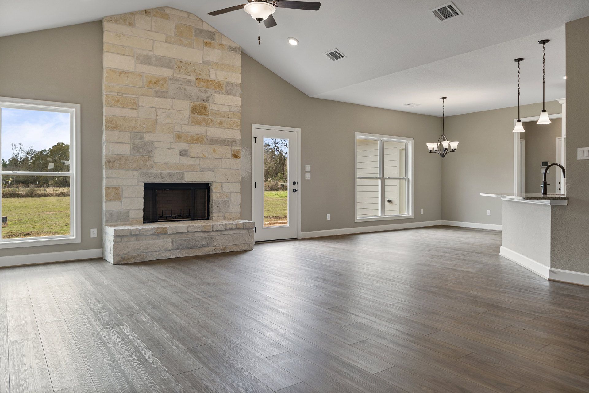 Spacious living room featuring hardwood floors, central stone fireplace with black frame, white-framed windows overlooking trees, and door opening to a field and wooded landscape.