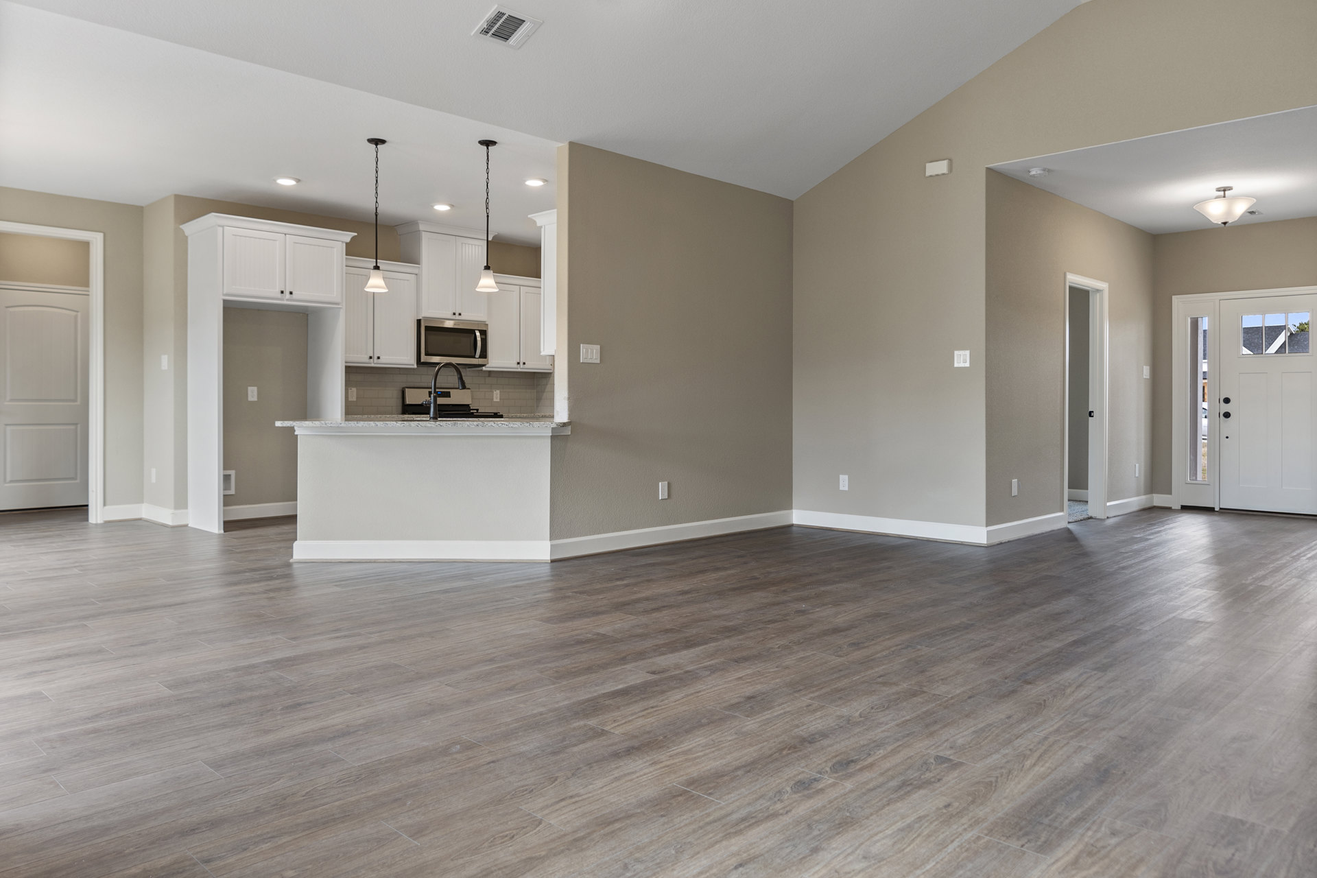 Spacious open floor plan featuring wood laminate flooring, modern kitchen with open microwave, adjacent dining area, white framed doors including one with a window, smooth plaster