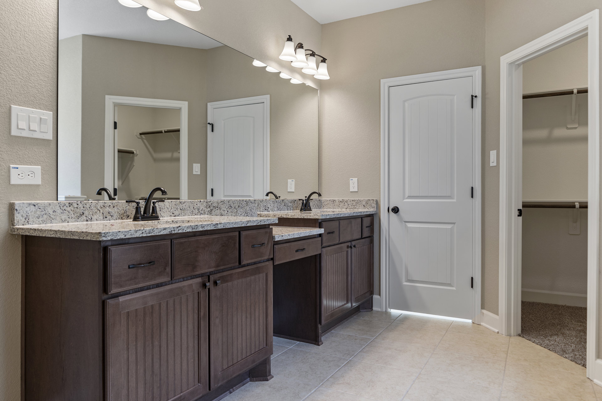 Bathroom with double sinks, white cabinetry, marble countertop, white door with black hardware, wall-mounted mirror with white frame, metal shelf, visible light switch and