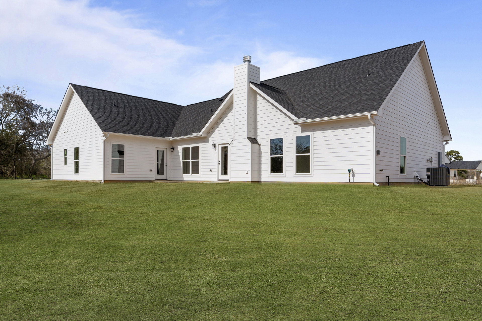 White farmhouse with gabled roof, brick chimney, large windows reflecting leafless tree, manicured green lawn, and cloudy sky backdrop