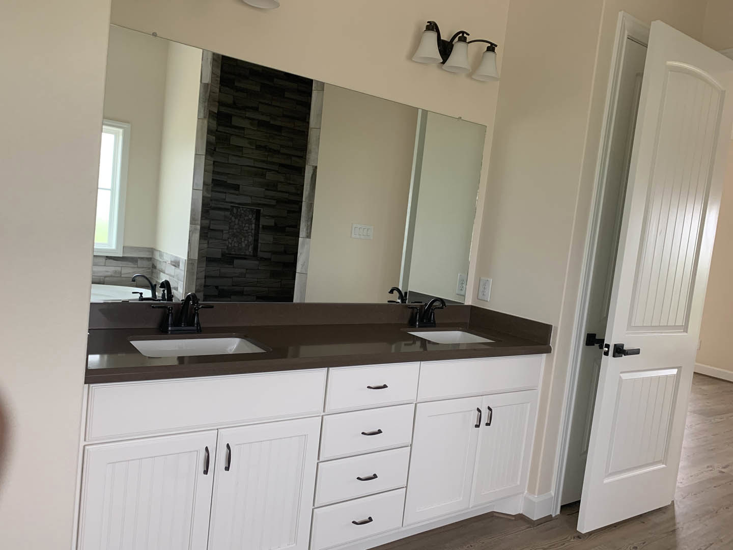 Bathroom with white shaker cabinets, wide framed mirror above quartz countertop, three-light fixture with white shades, black hardware on drawers and door, white tile backsplash