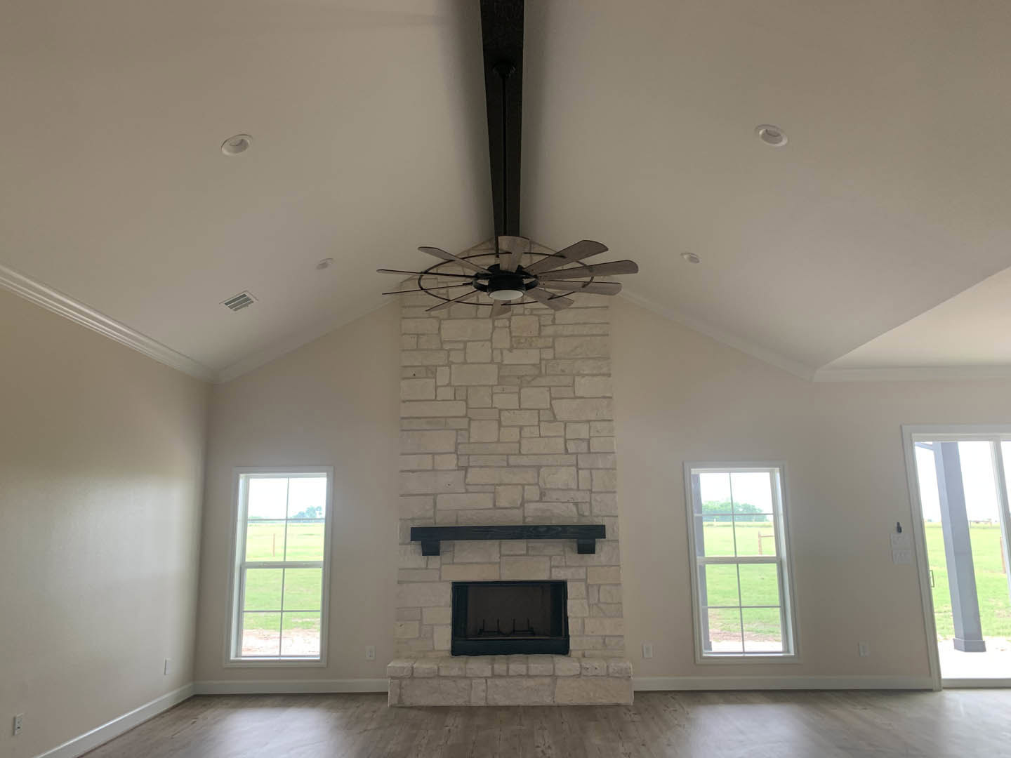 Living room with white walls, central fireplace with black hearth, ceiling fan with light, large windows overlooking grassy field, wood flooring, and decorative molding