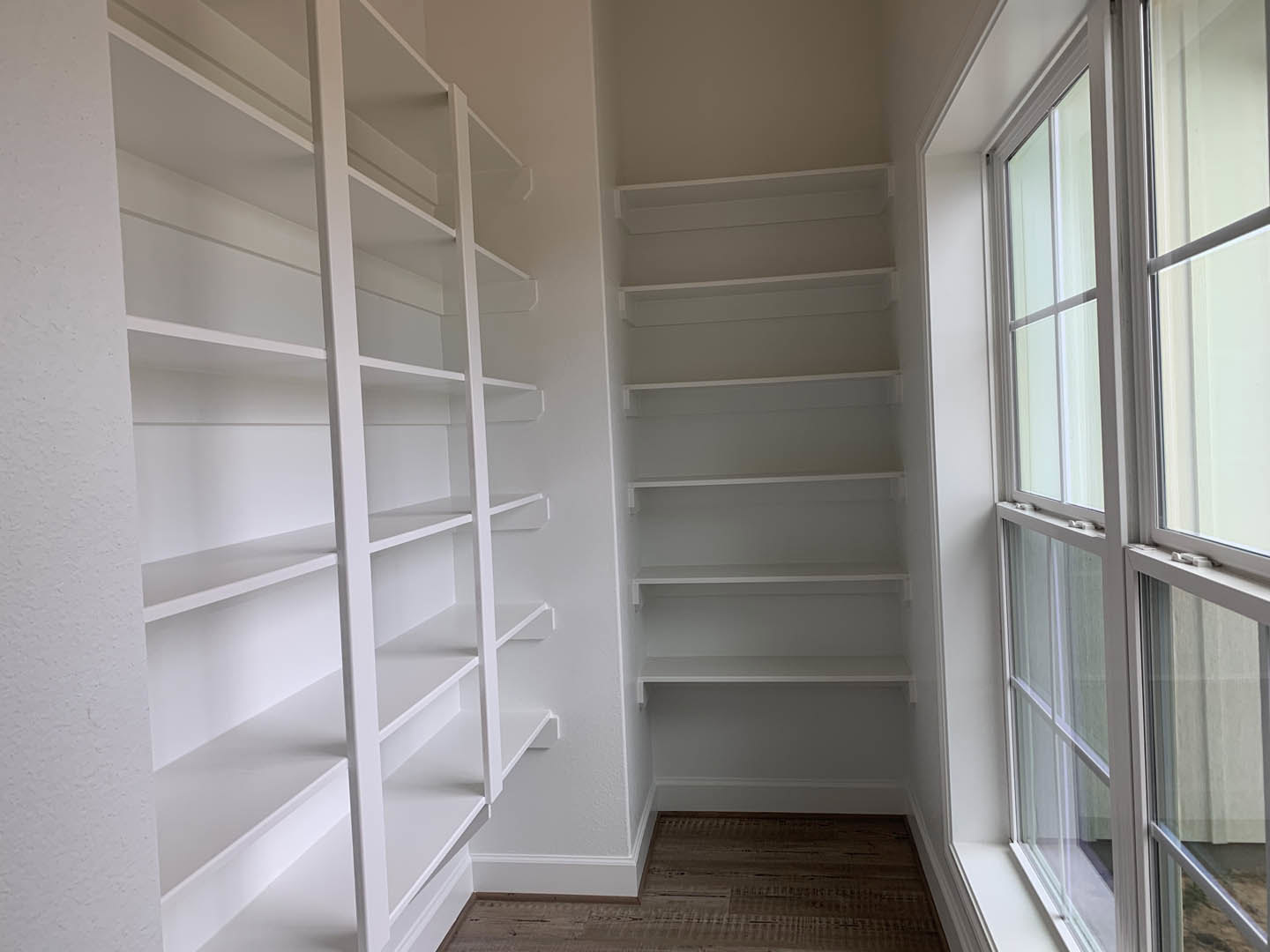 White built-in shelves along a wall, large window with glass panels, wood flooring, and white plaster walls in an empty room.