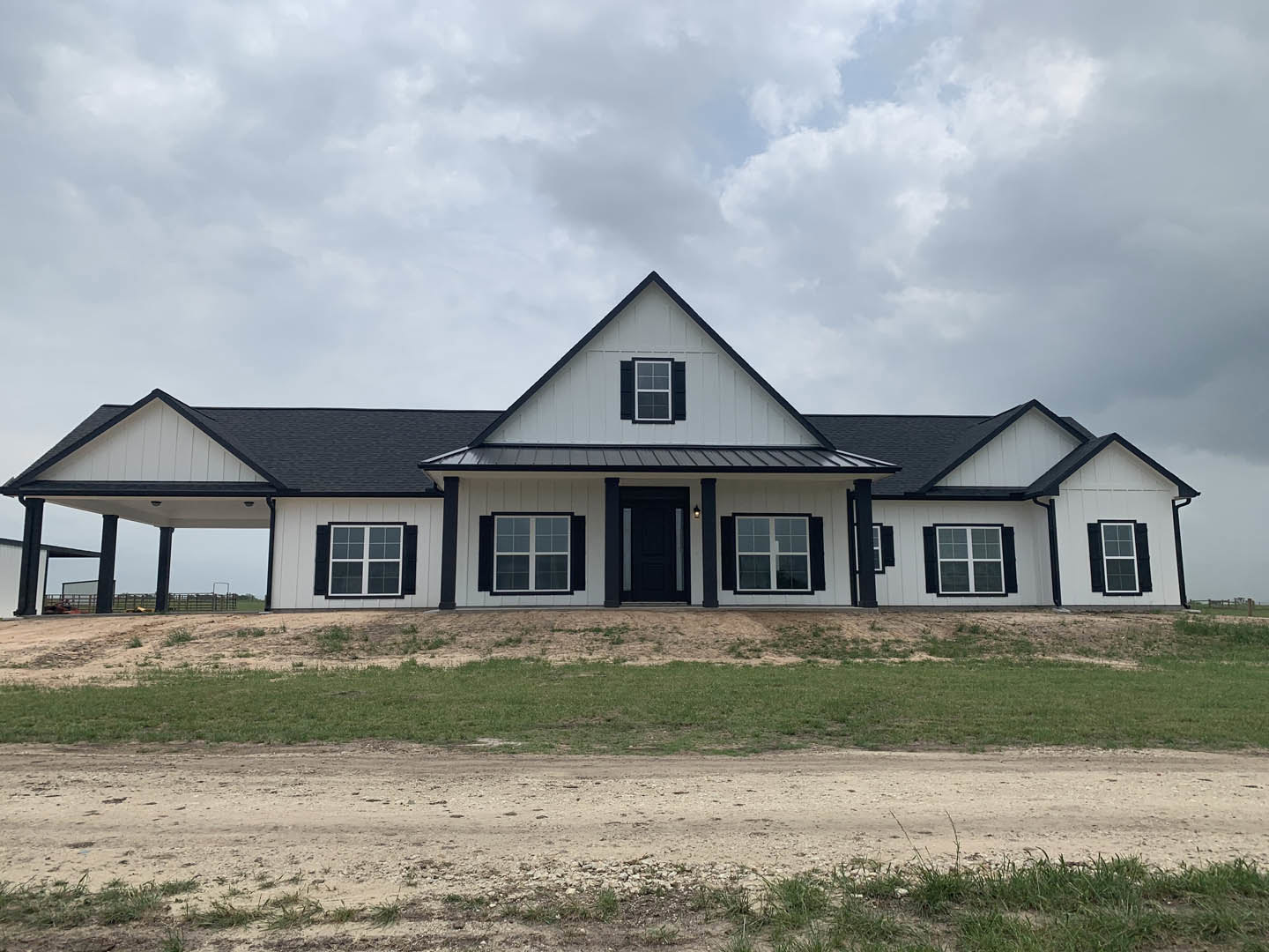 White siding house with black shingle roof, black door framed in white, black shuttered window, dirt driveway in foreground, blue sky overhead