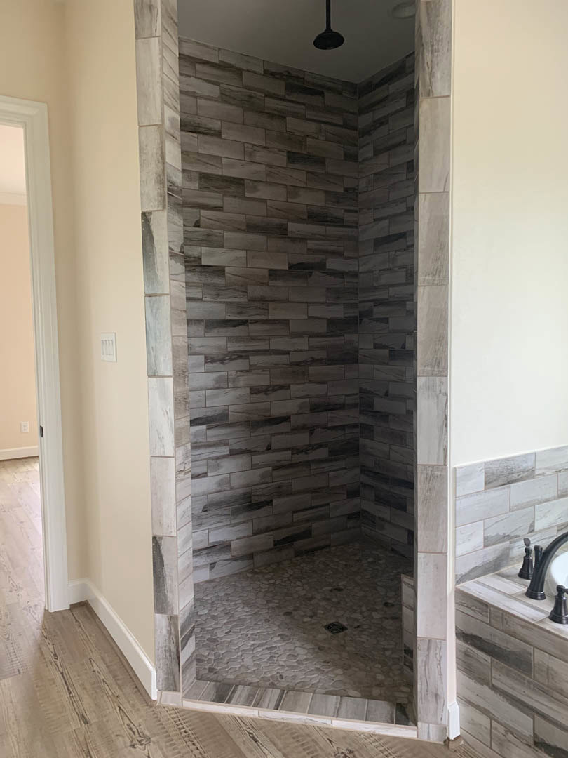 Walk-in shower with patterned tile wall, black shower fixture and pole, tiled floor with central drain, white door frame, pink painted wall, and plaster ceiling.
