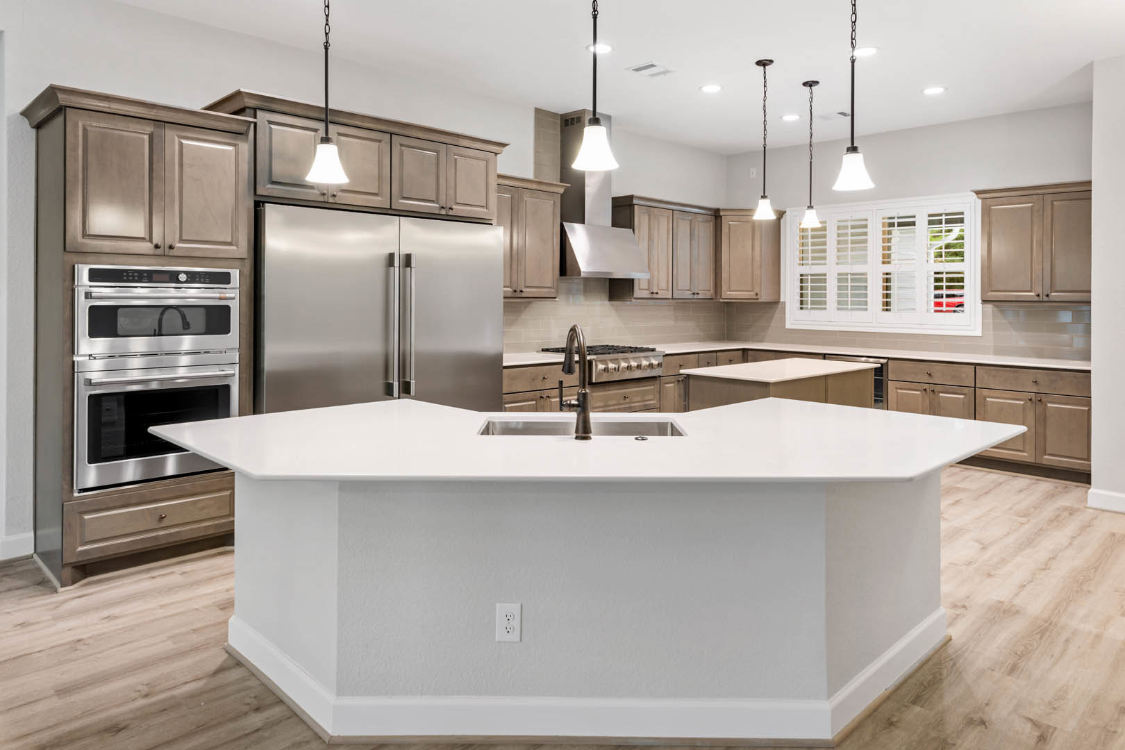 White kitchen with large central island featuring built-in sink, stainless steel oven, white cabinetry, stone countertops, refrigerator, window, and white electrical outlet.
