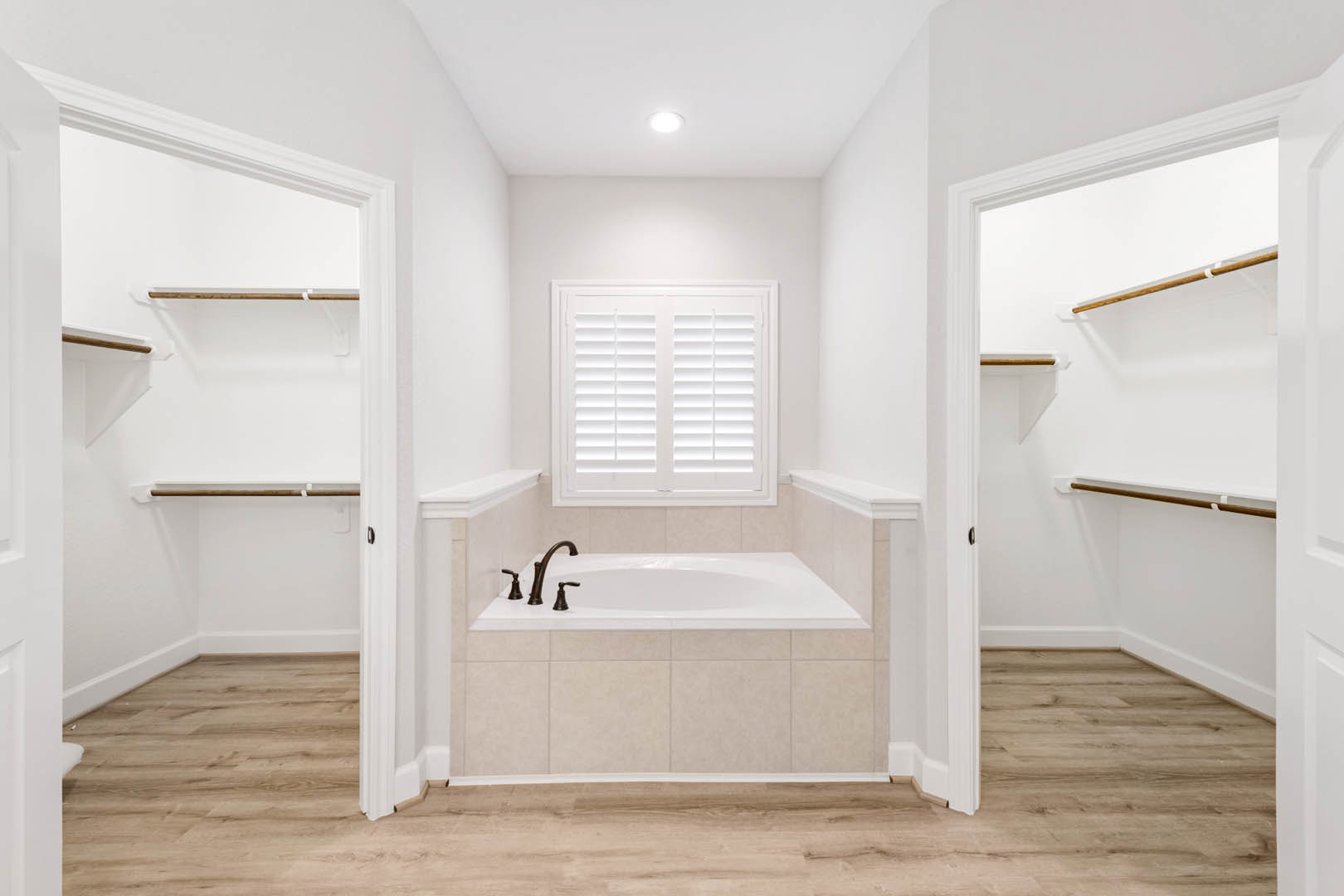 Freestanding bathtub beside white tile wall, wooden floor, built-in closets, chrome faucet, window, and floating wooden shelf