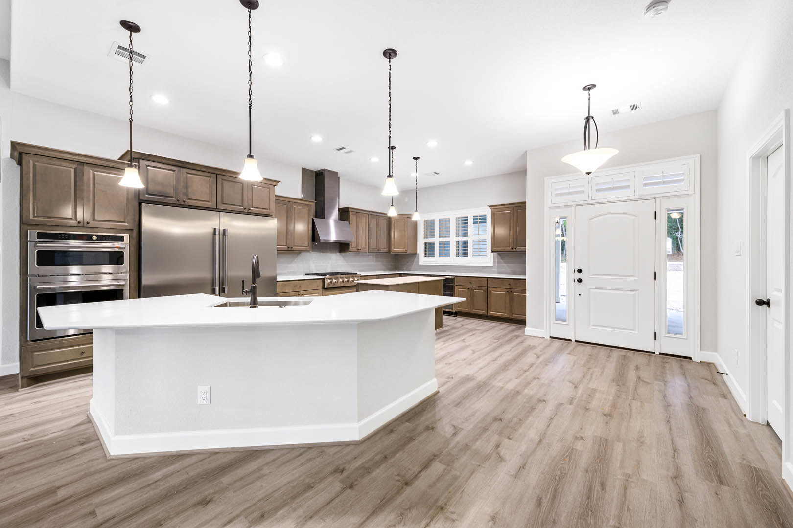 Spacious kitchen featuring a large white island with built-in sink, wood flooring, stainless steel oven and refrigerator, white cabinetry, and a glass-paneled door with black