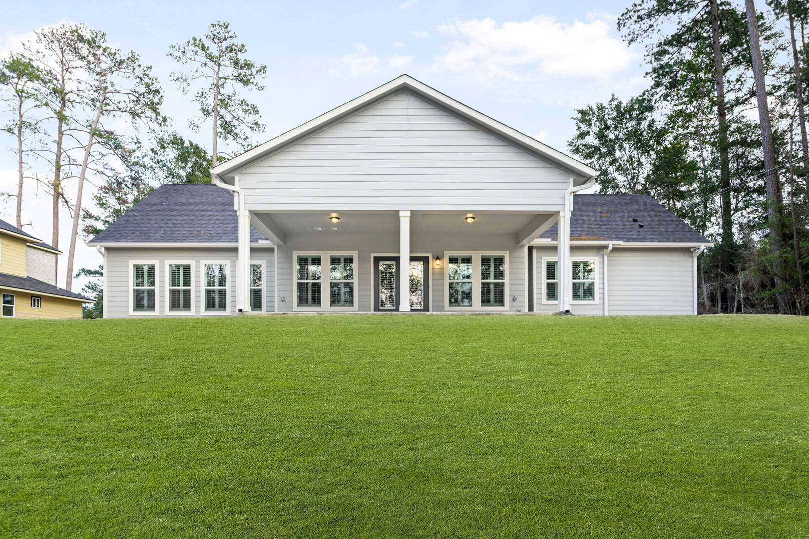Two-story custom home with white siding, large covered porch, multiple windows with white trim, expansive green lawn, mature trees, and blue sky in background
